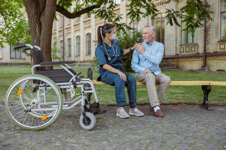 Full length shot of young nurse wearing face shield supporting aged man, recovering male patient, sitting together on the bench in park near hospitalの写真素材