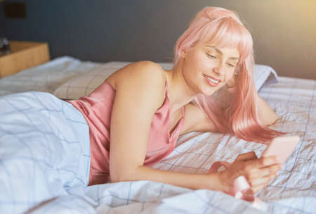 Pretty woman uses modern mobile phone lying on large bed in morningの写真素材