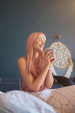 Pleasured lady with pink hair holds mug of delicious drink sitting on bedの写真素材