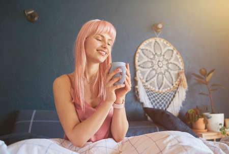 Pretty woman with pink hair holds mug sitting on comfortable bed with soft blanketの写真素材