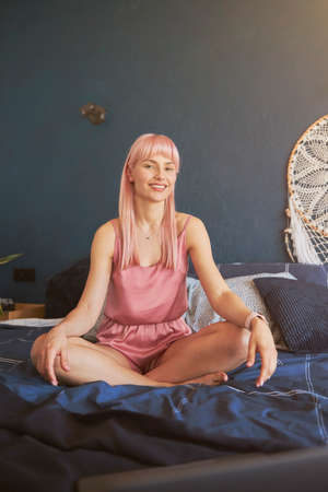 Female model with long pink hair in elegant pajamas sits in lotus pose on comfortable bedの写真素材