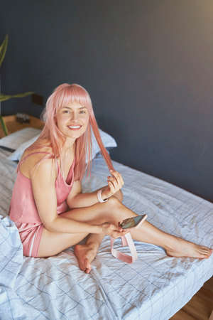 Positive person with messy pink hair holds smartphone sitting on bedの写真素材