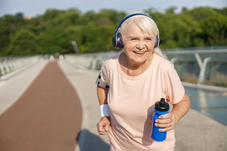 Positive senior lady with headphones runs along bridge in summerの写真素材
