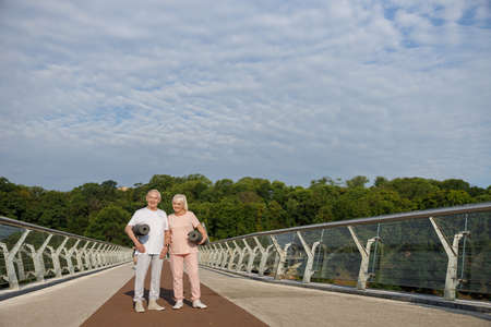 Positive senior couple with rolled mats and earphones stands on footbridgeの写真素材