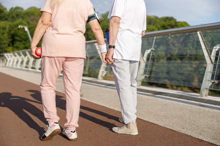 Senior couple in tracksuits walks joining hands along footbridge at sunsetの写真素材