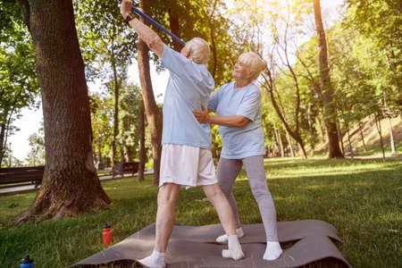 Positive mature woman supports back of senior man doing exercises with bar in parkの写真素材
