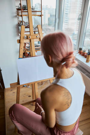 Woman with bun on head sits by easel on light balcony at homeの写真素材