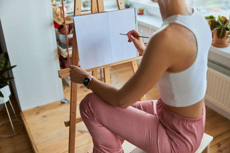 Young woman draws picture on canvas with brush at wooden easel on balconyの写真素材