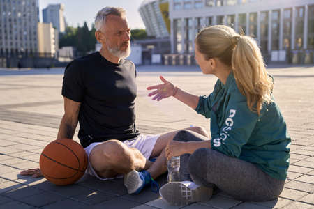 Middle aged couple, active man and woman in sportswear talking while relaxing after basketball workout outdoors on a sunny dayの写真素材