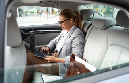 Working in taxi. Side view of a successful business woman wearing eyeglasses using laptop while sitting on back seat in the carの写真素材