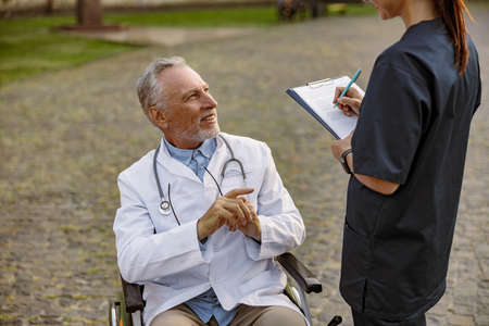 Smiling senior handicapped male doctor in wheelchair wearing lab coat having conversation with nurse making notes while assisting him outdoorsの写真素材