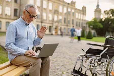 Senior man, recovering patient with wheelchair having video call, working on a laptop while sitting on the bench in the park near clinicの写真素材