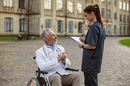 Successful senior handicapped male doctor in wheelchair wearing lab coat talking to young nurse making notes while assisting him outdoorsの写真素材