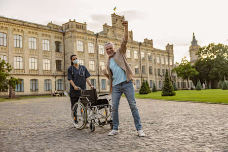 Joyful disabled handicapped mature man can walk again. Recovering patient standing up from his wheelchair, young nurse assisting him outdoorsの写真素材