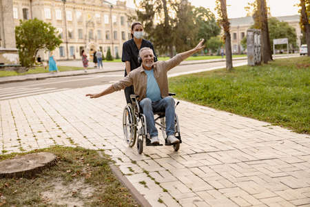 Excited mature disabled man in wheelchair wearing headphones having fun during a walk in the city assisted by young nurse in protective face maskの写真素材