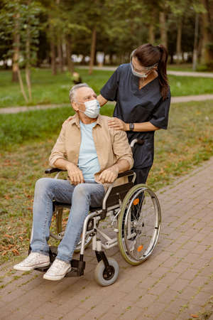 Young nurse in face shield and protective face mask taking care of mature handicapped man in wheelchair during a walk in the parkの写真素材