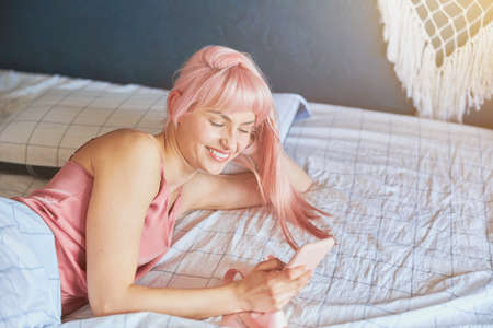 Cheerful lady with surfs internet with modern mobile phone in comfortable bedの写真素材