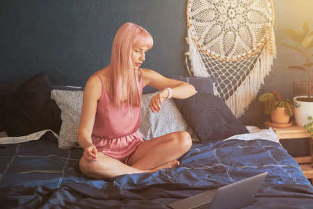 Worried lady with long pink hair checks time on smartwatch sitting on large bedの写真素材