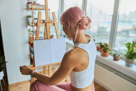 Elegant woman artist sits by easel with blank white canvas on at homeの写真素材