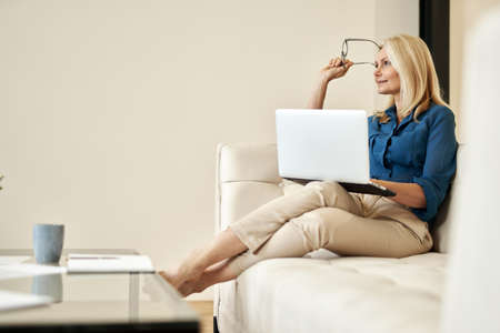 Pensive mature businesswoman smiling away while working from home, sitting on a couch in modern apartmentの写真素材