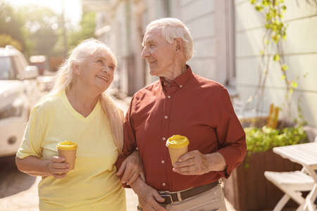 Positive senior family couple with cups walks together along streetの写真素材