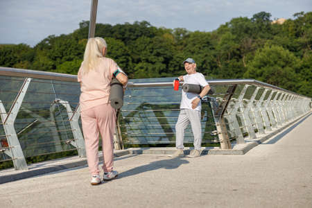 Smiling senior man with mat greets walking woman on footbridgeの写真素材