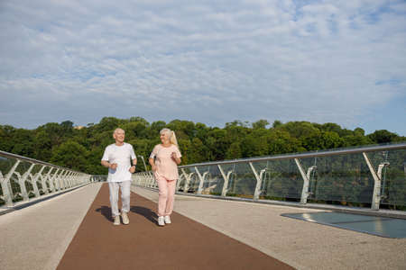 Smiling senior man and lady with mobile phone run along footbridgeの写真素材