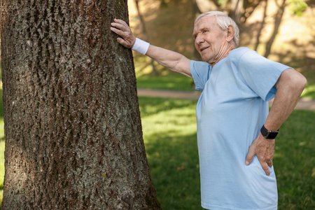 Positive senior sportsman rests leaning onto tree trunk at training in parkの写真素材