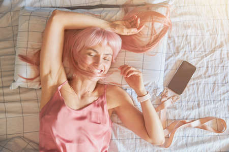 Relaxed woman holds lock of pink hair above face lying near phone on bedの写真素材