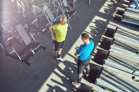 Middle aged man in sportswear warming up, stretching his legs and talking with fitness instructor at gym or sport club, full length shotの写真素材