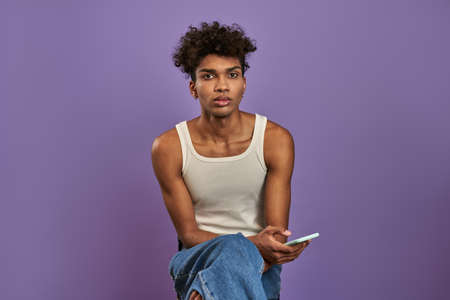 Close-up portrait of young man sits with mobile and looking camera on isolated purple backgroundの写真素材