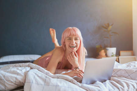 Attractive woman with bright pink laughs using modern laptop on bed by blue wallの写真素材