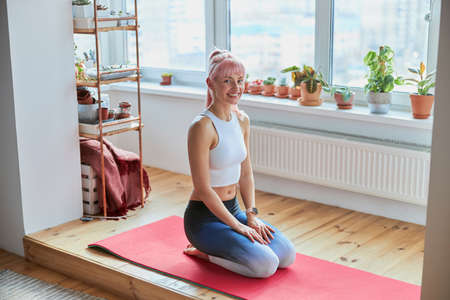 Woman with pink hair in tracksuit sits on knees at home trainingの写真素材
