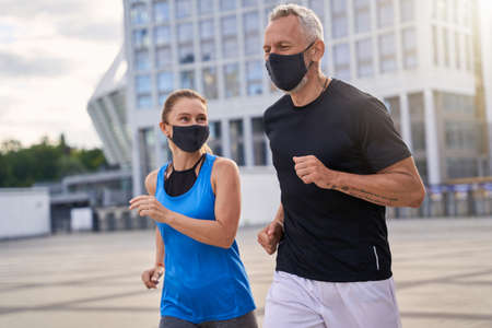 Active middle aged couple, woman and man wearing protective masks while running outdoors in the city to stay fit during Covid19 pandemicの写真素材