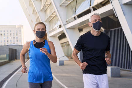 Cheerful middle aged couple wearing protective face masks running together in urban environment during coronavirus outbreakの写真素材