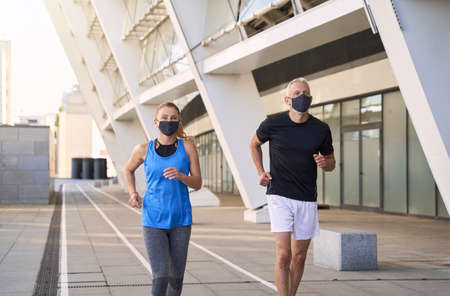 Active mature couple wearing protective face masks jogging together in urban environmentの写真素材