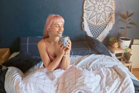 Beautiful lady holds cup sitting on comfortable bed in lazy morningの写真素材