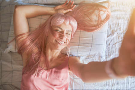 Joyful lady with bright pink hair takes selfie lying on comfortable bedの写真素材