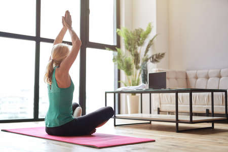 Sportive mature blonde woman practicing yoga, sitting in lotus position on the floor and watching online training using laptop at homeの写真素材