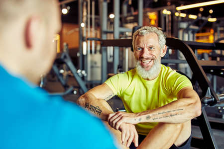 Portrait of a happy middle aged man discussing training results with fitness instructor or personal trainer and smiling while sitting together on the floor at gymの写真素材