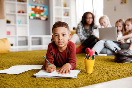 Smiling little boy holding a pencil and drawing on paper on the floorの写真素材