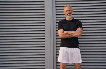 Attractive middle aged man in perfect shape smiling at camera while standing with arms crossed outdoors after workoutの写真素材