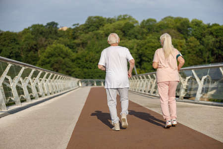 Sportive senior man and woman in tracksuits run together along footbridgeの写真素材
