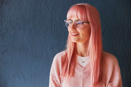 Beautiful woman with pink hair and glasses wearing sweater stands near blue wallの写真素材