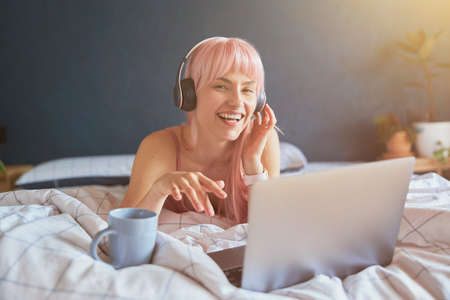 Joyful woman with headphones listens to music on laptop on bedの写真素材