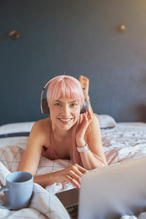 Stylish lady with headphones and cup of drink watches video via laptop on bedの写真素材
