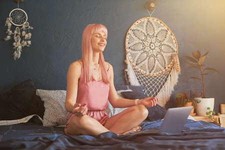 Pretty smiling female model meditates near laptop on large bed by dark blue wallの写真素材