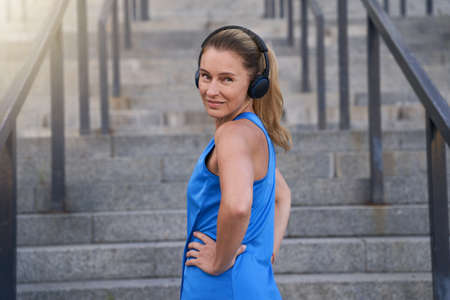 Active sportswoman in headphones looking at camera, standing with hands on the waist outdoors ready for training in the cityの写真素材