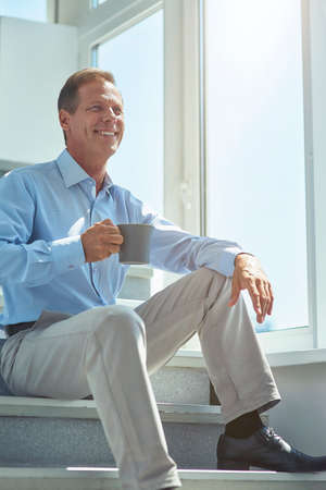 Taking a break at work. Vertical shot of a happy mature businessman in classic wear sitting on stairs in the office and drinking fresh coffeeの写真素材
