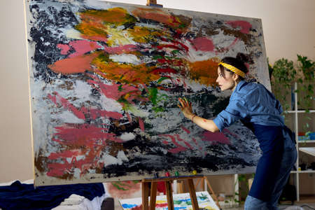 Creative young female painter working on a large modern abstract oil painting, applying paint on canvas with fingers in living room at home studio workshopの写真素材